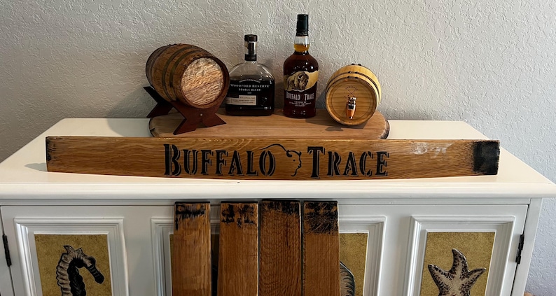 May include: A wooden sign with the text "BUFFALO TRACE" and a buffalo head logo. The sign is displayed on a white cabinet, with two small wooden barrels and two bottles of liquor on top. The barrels are made of wood and have a tap.