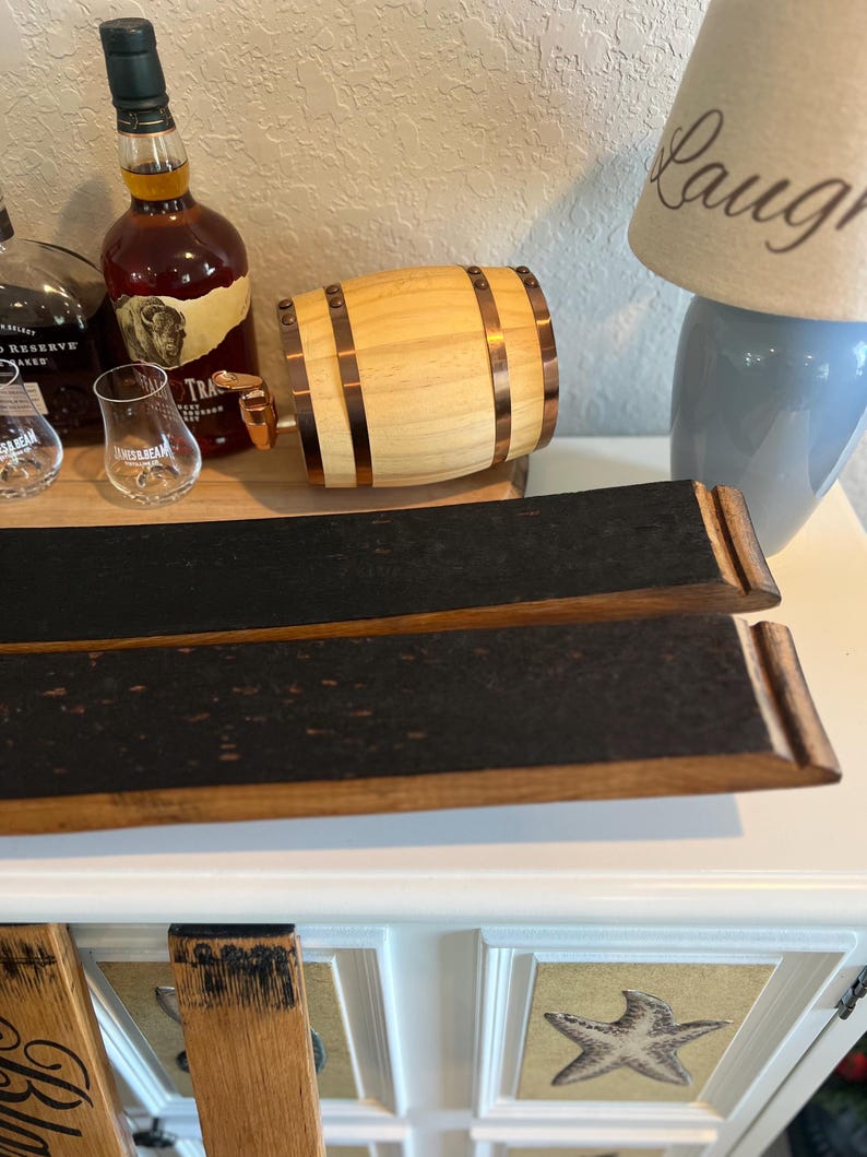 May include: A wooden barrel-shaped dispenser with copper-colored bands and a spigot. Two curved, charred wood pieces are in the foreground. A bottle of liquor and two glasses are on a wooden tray. A lamp with the word "Laugh" is in the background.