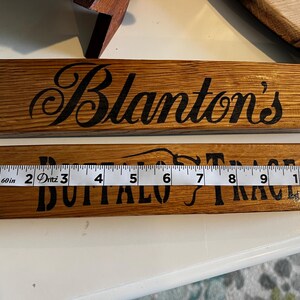 Puede incluir: Dos letreros de madera con letras negras. El letrero superior dice "Blanton's" en una fuente cursiva. El letrero inferior dice "Buffalo Trace". Una cinta m&eacute;trica muestra que el letrero mide aproximadamente 28 cm de largo.