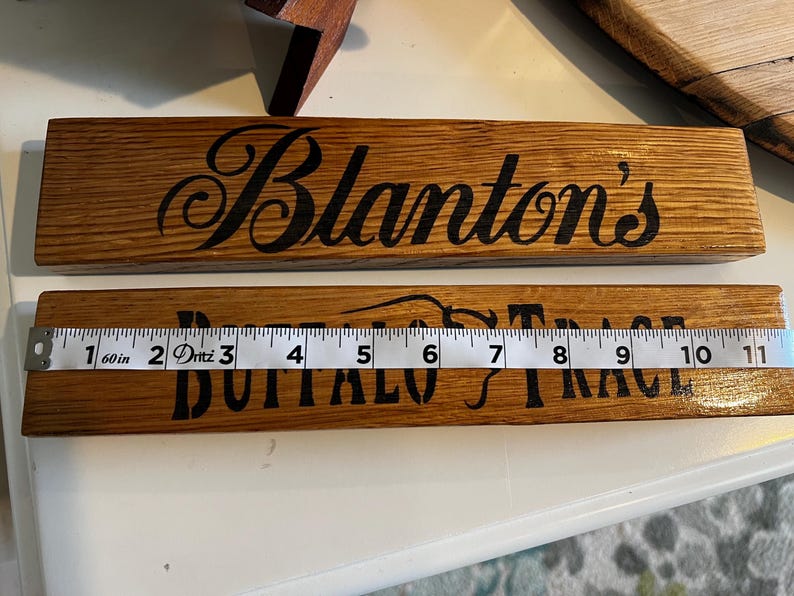 Puede incluir: Dos letreros de madera con letras negras. El letrero superior dice "Blanton's" en una fuente cursiva. El letrero inferior dice "Buffalo Trace". Una cinta m&eacute;trica muestra que el letrero mide aproximadamente 28 cm de largo.