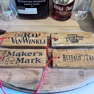 May include: Four wooden bourbon barrel stave tags with red string. The tags are branded with the names "Old Rip Van Winkle", "Maker's Mark", "Kentucky Bourbon Trail", and "Buffalo Trace". Bottles and glasses are in the background.