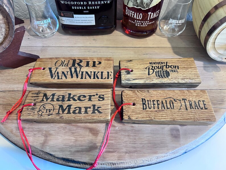 May include: Four wooden bourbon barrel stave tags with red string. The tags are branded with the names "Old Rip Van Winkle", "Maker's Mark", "Kentucky Bourbon Trail", and "Buffalo Trace". Bottles and glasses are in the background.