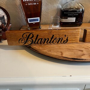 Puede incluir: Un letrero de madera con el nombre "Blanton's" en escritura negra. El letrero est&aacute; hecho de madera de color marr&oacute;n claro y se muestra con dos botellas de bourbon y una copa de degustaci&oacute;n.