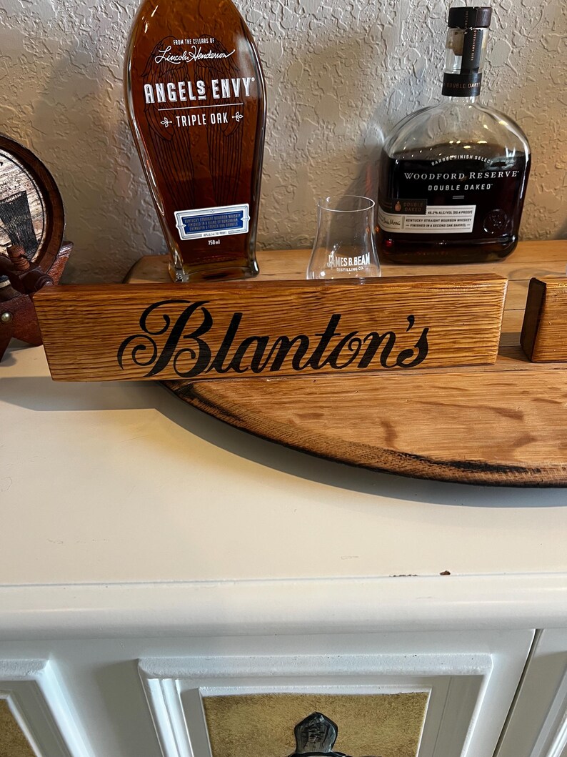 Puede incluir: Un letrero de madera con el nombre "Blanton's" en escritura negra. El letrero est&aacute; hecho de madera de color marr&oacute;n claro y se muestra con dos botellas de bourbon y una copa de degustaci&oacute;n.
