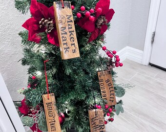 May include: A decorated Christmas tree with wooden ornaments. The ornaments are rectangular and feature the names "Maker's Mark", "Buffalo Trace", "Bullet Bourbon", and "Rip Van Winkle". The tree is adorned with red poinsettias and berries.