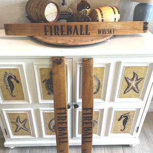 Puede incluir: Letrero de madera con el texto "FIREBALL WHISKY" en letras negras. El letrero est&aacute; hecho de madera de color marr&oacute;n claro y se muestra con dos piezas verticales a juego. El fondo incluye botellas de whisky y un mueble decorativo.