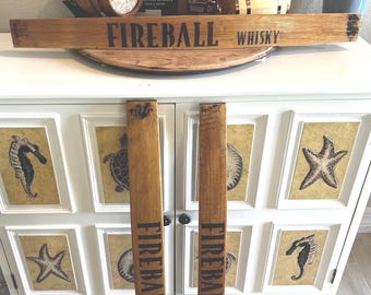May include: Wooden sign with the text "FIREBALL WHISKY" in black lettering. The sign is made of light brown wood and is displayed with two matching vertical pieces. The background includes bottles of whiskey and a decorative cabinet.