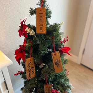 May include: A small Christmas tree decorated with wooden ornaments. The ornaments are rectangular and feature the names "Buffalo Trace", "Old Rip Van Winkle", "Makers Mark", and "Woodford Reserve Bourbon". The ornaments are suspended by red strings.