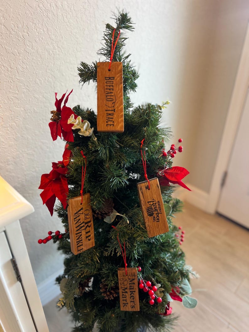 May include: A small Christmas tree decorated with wooden ornaments. The ornaments are rectangular and feature the names "Buffalo Trace", "Old Rip Van Winkle", "Makers Mark", and "Woodford Reserve Bourbon". The ornaments are suspended by red strings.