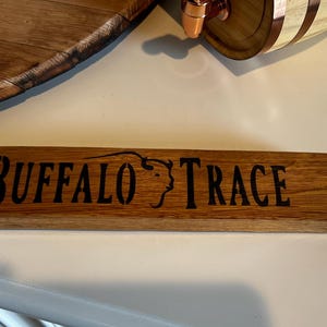 Puede incluir: Un letrero rectangular de madera con las palabras "BUFFALO TRACE" en negro y la silueta de una cabeza de b&uacute;falo. El letrero es de madera de color marr&oacute;n claro. Ideal para un bar o destiler&iacute;a.