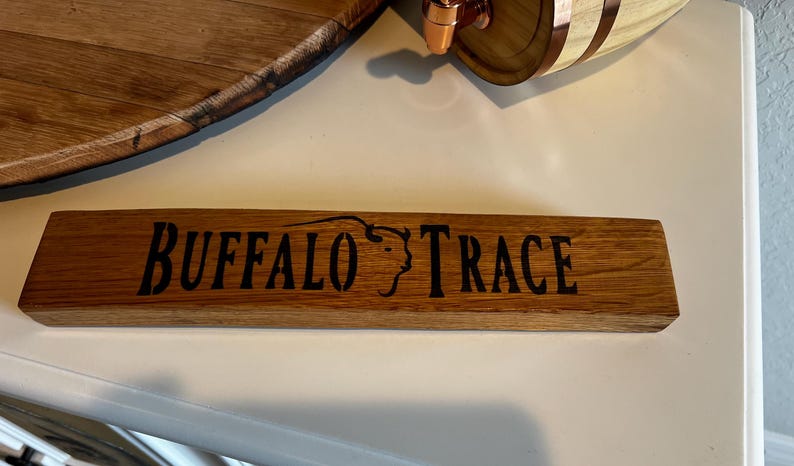 Puede incluir: Un letrero rectangular de madera con las palabras "BUFFALO TRACE" en negro y la silueta de una cabeza de b&uacute;falo. El letrero es de madera de color marr&oacute;n claro. Ideal para un bar o destiler&iacute;a.