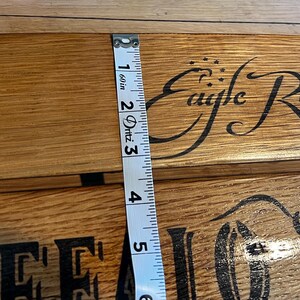 Op de afbeelding: Close-up van een houten oppervlak met de woorden "Eagle Rare" in een decoratief lettertype. Een meetlint toont een breedte van ongeveer 12,7 cm. Het hout heeft een natuurlijke nerf en een warme, bruine tint.