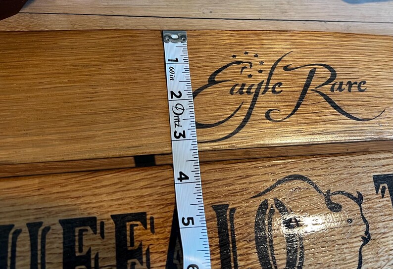 Op de afbeelding: Close-up van een houten oppervlak met de woorden "Eagle Rare" in een decoratief lettertype. Een meetlint toont een breedte van ongeveer 12,7 cm. Het hout heeft een natuurlijke nerf en een warme, bruine tint.