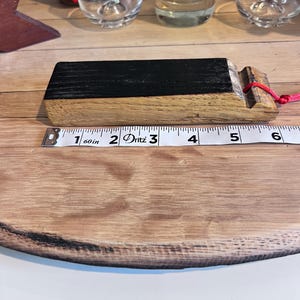 May include: A wooden bottle stopper with a black top and a red string. The stopper is approximately 5 inches long. The stopper is on a wooden surface with a measuring tape.
