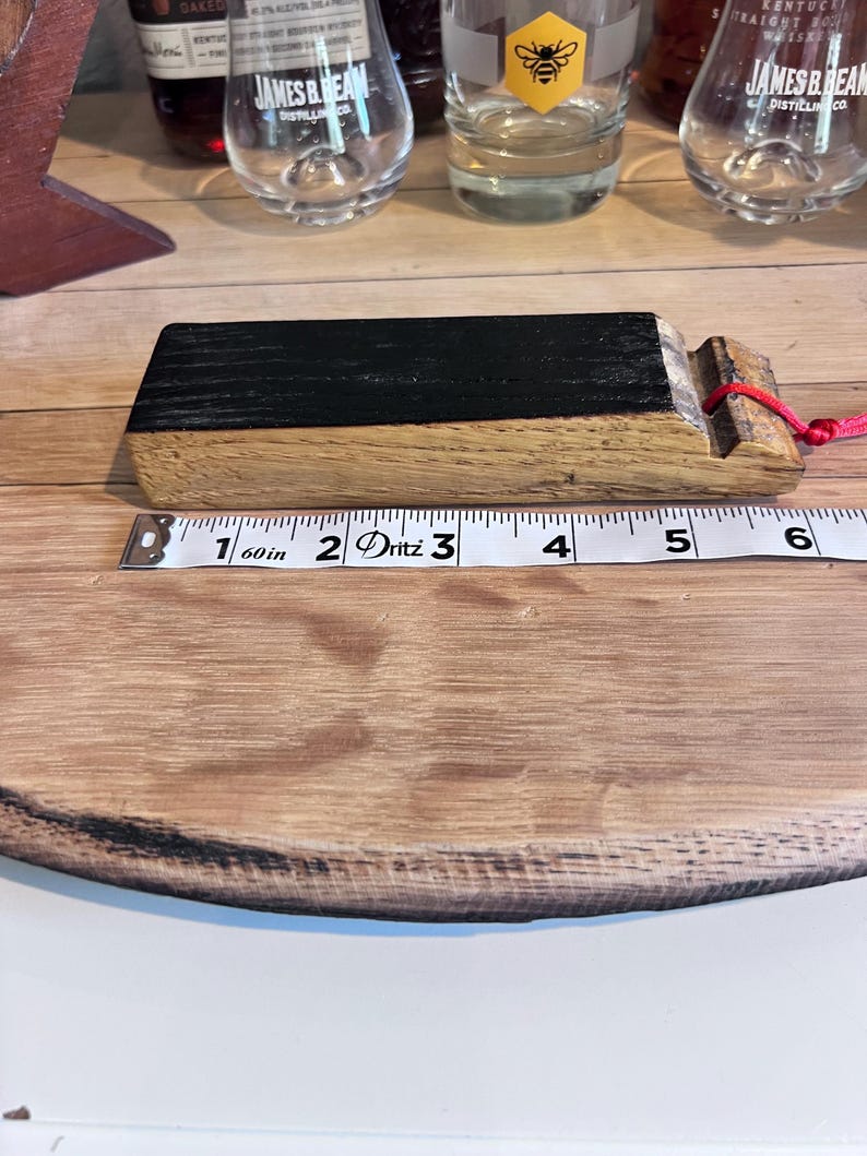 May include: A wooden bottle stopper with a black top and a red string. The stopper is approximately 5 inches long. The stopper is on a wooden surface with a measuring tape.