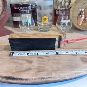May include: A wooden barrel bung with a black and brown finish, attached to a red string. The bung measures approximately 3 inches in length. Whiskey glasses and a barrel are in the background.