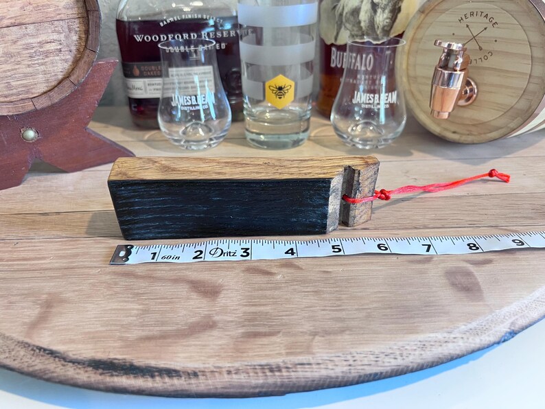 May include: A wooden barrel bung with a black and brown finish, attached to a red string. The bung measures approximately 3 inches in length. Whiskey glasses and a barrel are in the background.
