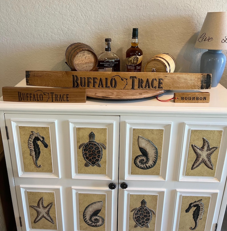 Puede incluir: Un mueble blanco con arte enmarcado de criaturas marinas, coronado con una decoraci&oacute;n tem&aacute;tica de bourbon Buffalo Trace. Incluye letreros de madera con texto "Buffalo Trace" y "Bourbon", botellas de bourbon y peque&ntilde;os barriles de madera.
