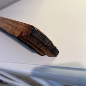 May include: Close-up of a wooden object with a dark brown, textured surface. The object appears to be a piece of a larger structure, possibly a frame or a decorative element. The wood grain is visible, and the edges are rough.