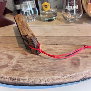 May include: A handcrafted wooden bottle stopper with a rectangular shape and a red cord attached. The stopper is made from a dark, textured wood and is displayed on a wooden surface. Several glasses and bottles are visible in the background.
