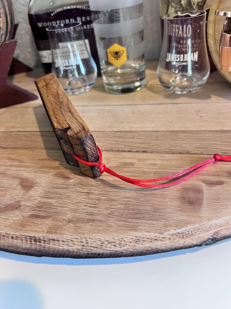 May include: A handcrafted wooden bottle stopper with a rectangular shape and a red cord attached. The stopper is made from a dark, textured wood and is displayed on a wooden surface. Several glasses and bottles are visible in the background.