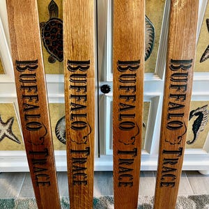 May include: Four wooden planks with the words "BUFFALO TRACE" in black lettering. The planks are a medium brown color with visible wood grain. The planks are leaning against a white cabinet with decorative elements.