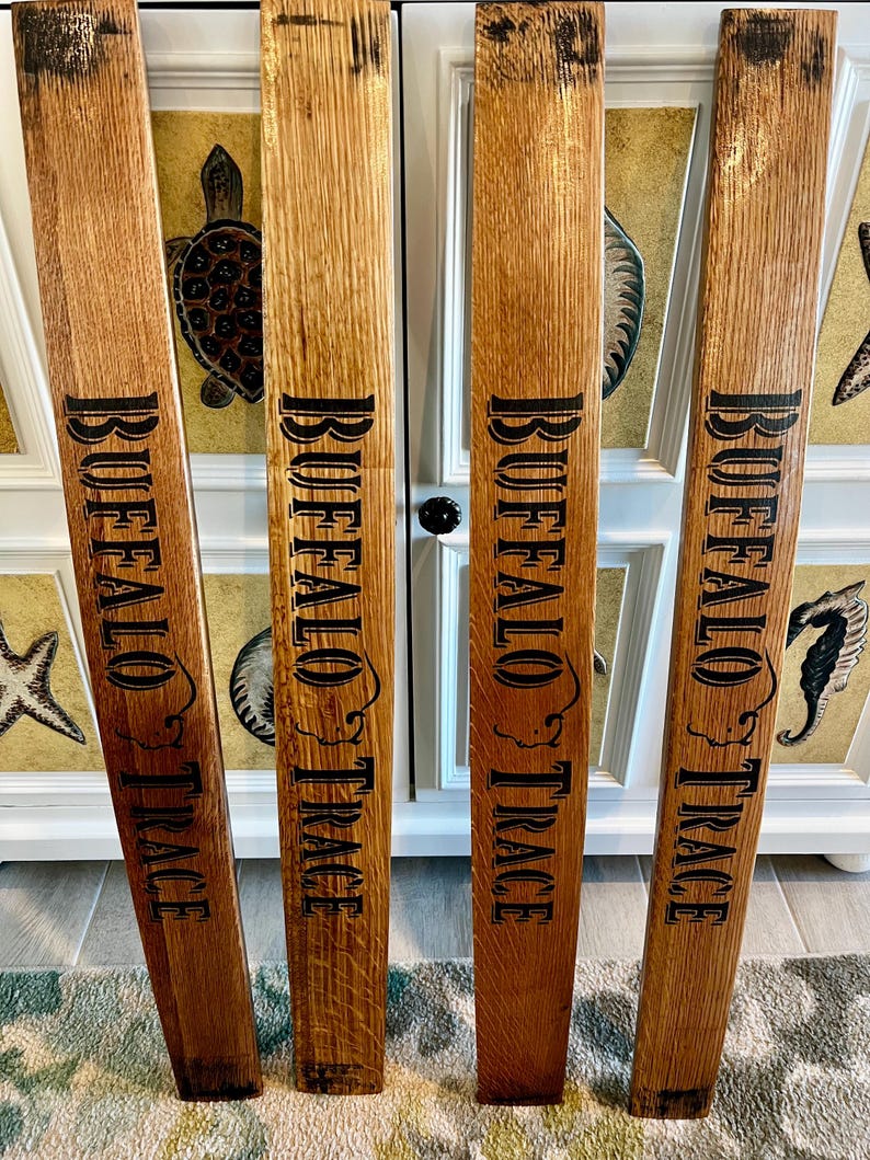 May include: Four wooden planks with the words "BUFFALO TRACE" in black lettering. The planks are a medium brown color with visible wood grain. The planks are leaning against a white cabinet with decorative elements.