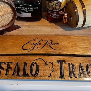 Op de afbeelding: Houten vatenplank met de woorden "Buffalo Trace" en "Eagle Rare" in zwarte letters. Twee kleine houten vaten en twee whiskyflessen zijn zichtbaar op de achtergrond.