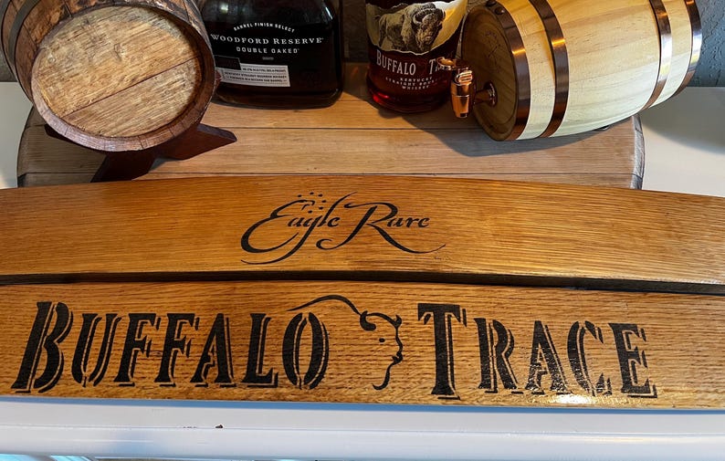 Op de afbeelding: Houten vatenplank met de woorden "Buffalo Trace" en "Eagle Rare" in zwarte letters. Twee kleine houten vaten en twee whiskyflessen zijn zichtbaar op de achtergrond.