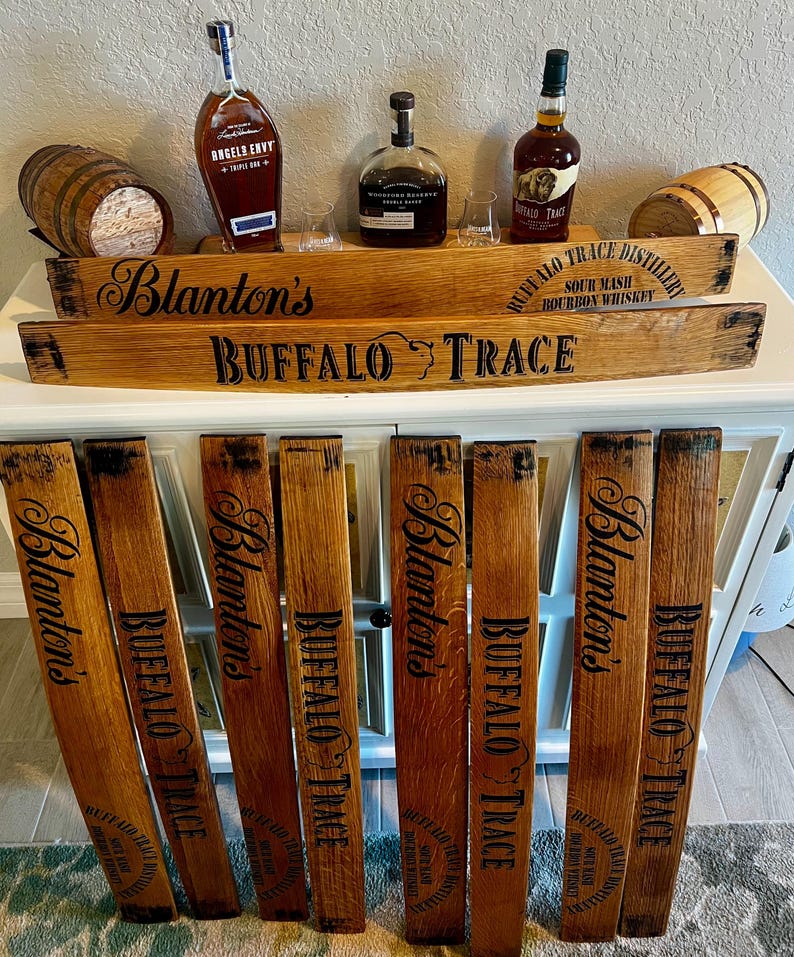 May include: A display of bourbon bottles and barrel stave signs. The signs are made of wood and feature the names "Blanton's" and "Buffalo Trace" in black lettering. The bottles are various shapes and sizes, with labels visible. Small wooden barrels are also present.