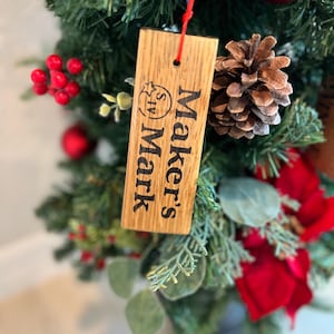 May include: A wooden Christmas tree ornament with the words "Maker's Mark" printed in black. The ornament is hanging from a red string and surrounded by green pine needles, red berries, and a pine cone.
