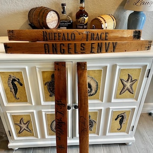 May include: Wooden barrel stave segments with the text "BUFFALO TRACE" and "ANGELS ENVY" in black lettering. Two additional staves are inscribed with names. The items are displayed with bottles of bourbon and small wooden barrels.