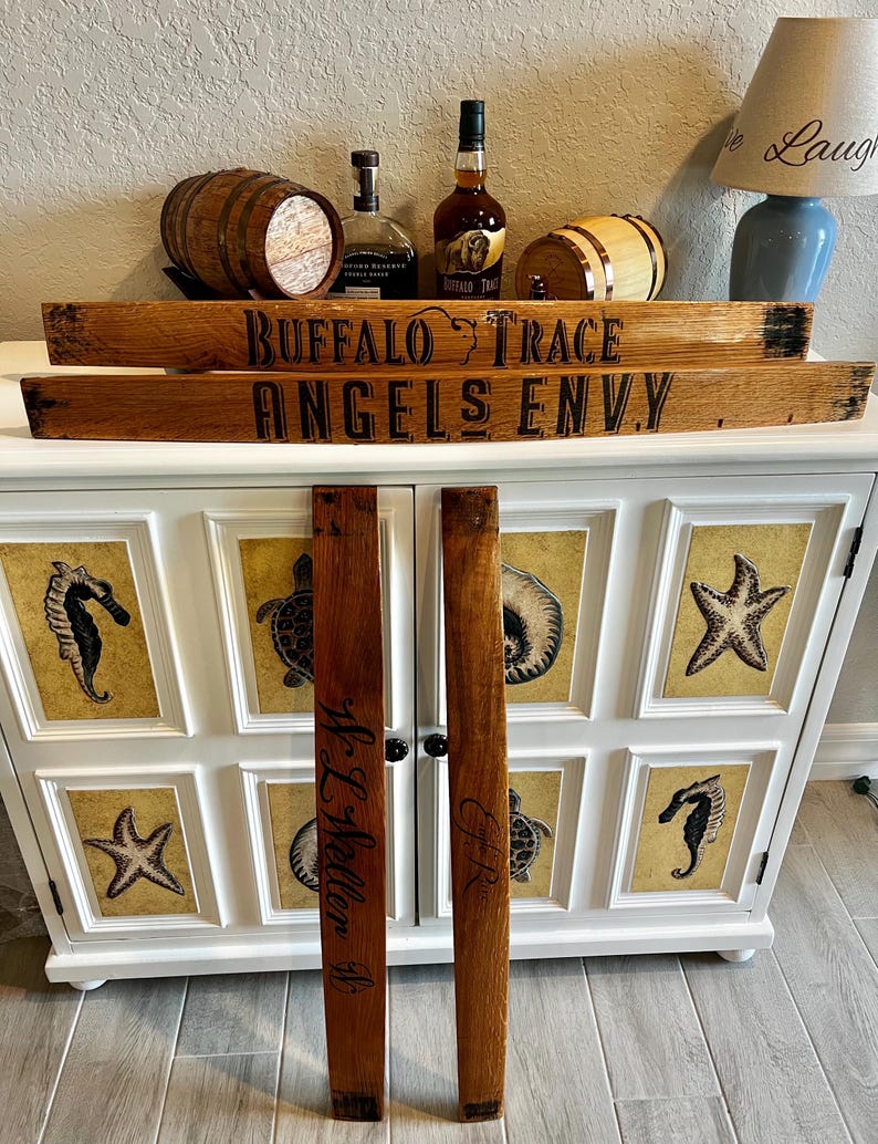 May include: Wooden barrel stave segments with the text "BUFFALO TRACE" and "ANGELS ENVY" in black lettering. Two additional staves are inscribed with names. The items are displayed with bottles of bourbon and small wooden barrels.
