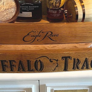 Op de afbeelding: Houten duigen met de tekst "Eagle Rare" en "Buffalo Trace" in zwart. De duigen worden weergegeven voor drankflessen en kleine houten vaten, wat wijst op een thema van whisky of bourbon.