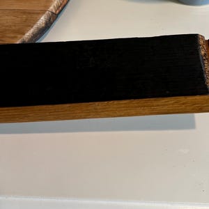 Puede incluir: Una secci&oacute;n de una duela de barril de madera con un centro rectangular negro y un borde de madera natural. El extremo de la duela tiene un detalle tallado. La veta de la madera es visible.