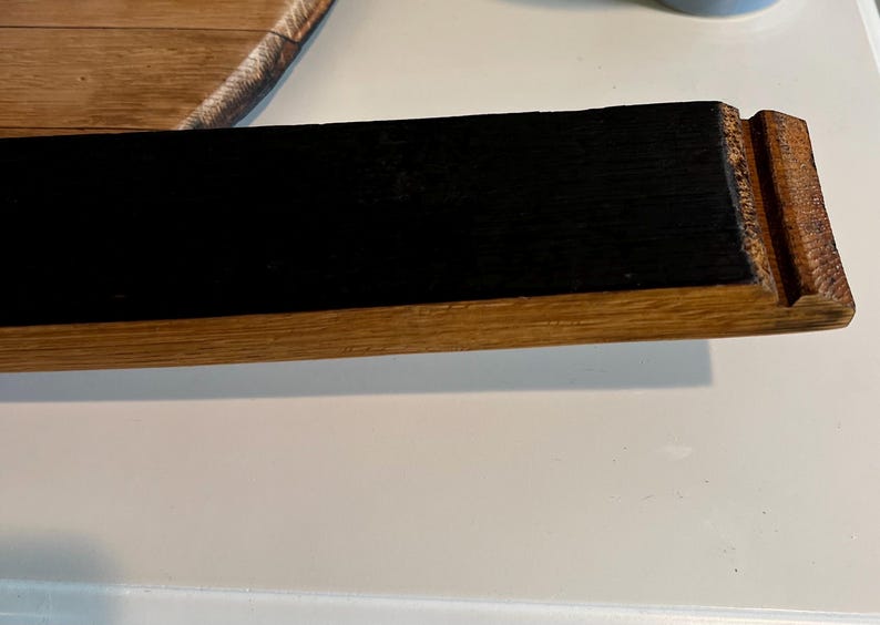 Puede incluir: Una secci&oacute;n de una duela de barril de madera con un centro rectangular negro y un borde de madera natural. El extremo de la duela tiene un detalle tallado. La veta de la madera es visible.