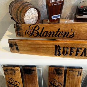 May include: Wooden barrel stave signs with the text "Blanton's" and "Buffalo". A small wooden barrel and bottles of Angels Envy and Woodford Reserve bourbon are also visible. The signs are made of light brown wood with black lettering.