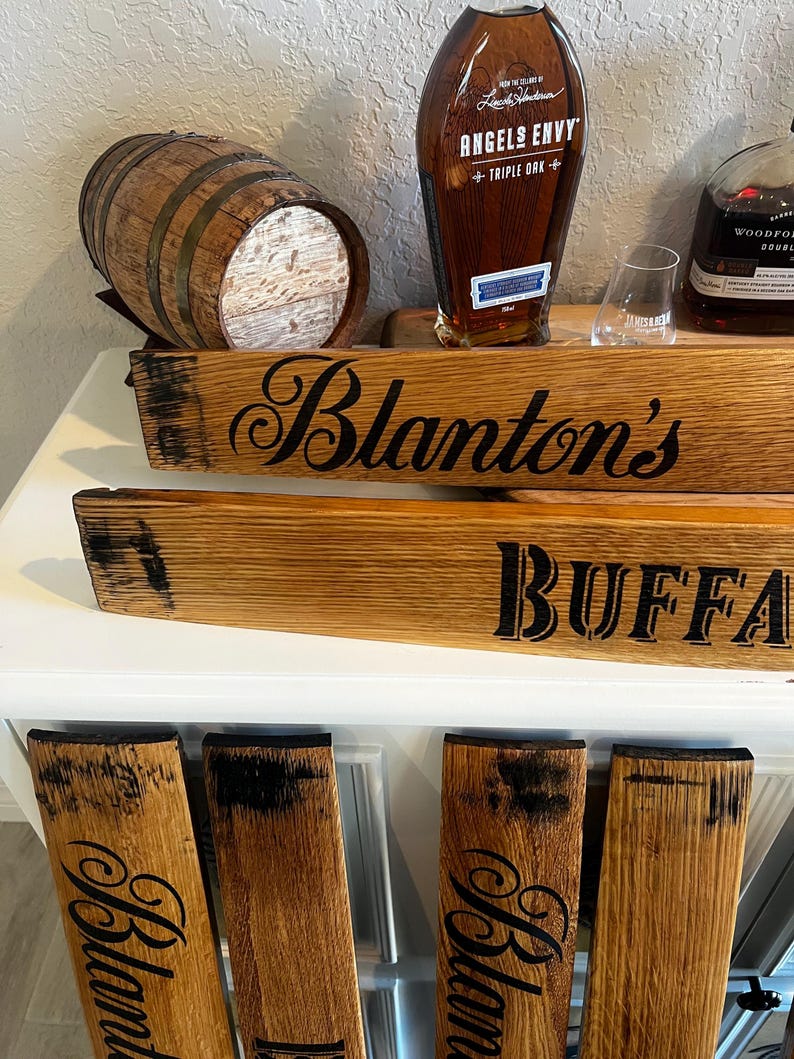 May include: Wooden barrel stave signs with the text "Blanton's" and "Buffalo". A small wooden barrel and bottles of Angels Envy and Woodford Reserve bourbon are also visible. The signs are made of light brown wood with black lettering.