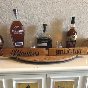 Puede incluir: Una exhibici&oacute;n de botellas de whisky y letreros de madera. Los letreros dicen "Blanton's" y "Buffalo Trace". Se ven botellas de whisky "Angel's Envy" y "Buffalo Trace". Tambi&eacute;n hay un peque&ntilde;o barril de madera.