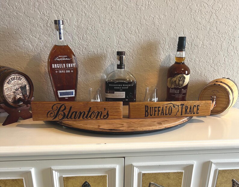 Puede incluir: Una exhibici&oacute;n de botellas de whisky y letreros de madera. Los letreros dicen "Blanton's" y "Buffalo Trace". Se ven botellas de whisky "Angel's Envy" y "Buffalo Trace". Tambi&eacute;n hay un peque&ntilde;o barril de madera.