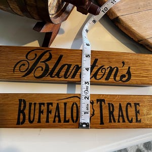 Puede incluir: Dos letreros rectangulares de madera con texto negro. El letrero superior dice "Blanton's" en cursiva. El letrero inferior dice "BUFFALO TRACE" en may&uacute;sculas. Los letreros miden aproximadamente 5 cm de ancho.