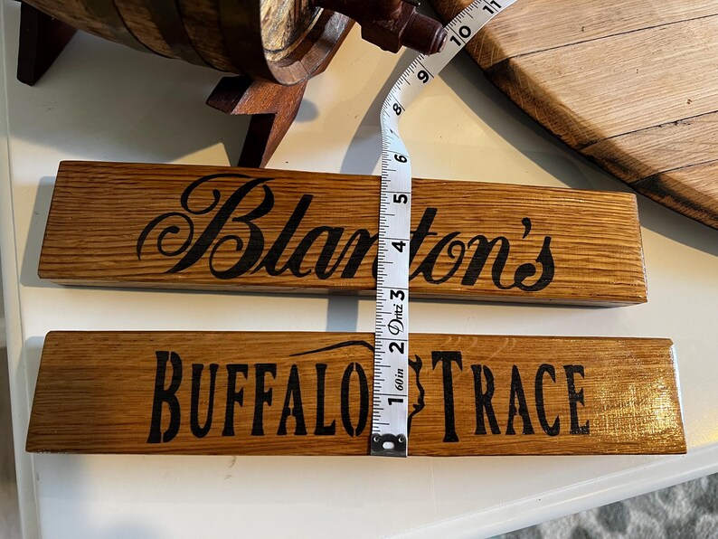 Puede incluir: Dos letreros rectangulares de madera con texto negro. El letrero superior dice "Blanton's" en cursiva. El letrero inferior dice "BUFFALO TRACE" en may&uacute;sculas. Los letreros miden aproximadamente 5 cm de ancho.
