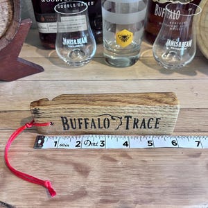 May include: A wooden Buffalo Trace whiskey barrel stave with the brand name printed in black. A red cord is attached. A measuring tape shows the stave is approximately 6 inches long. Whiskey glasses and bottles are in the background.