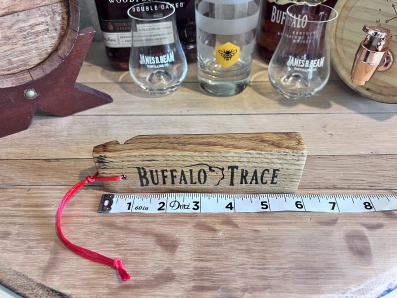 May include: A wooden Buffalo Trace whiskey barrel stave with the brand name printed in black. A red cord is attached. A measuring tape shows the stave is approximately 6 inches long. Whiskey glasses and bottles are in the background.