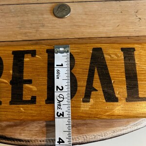 Puede incluir: Un letrero de madera con la palabra "REBAL" en letras negras. Una moneda de plata est&aacute; encima del letrero. Una cinta m&eacute;trica muestra que el letrero mide aproximadamente 7,6 cm de ancho.
