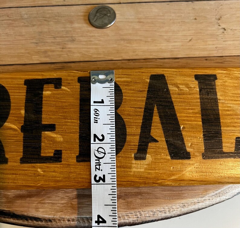 Puede incluir: Un letrero de madera con la palabra "REBAL" en letras negras. Una moneda de plata est&aacute; encima del letrero. Una cinta m&eacute;trica muestra que el letrero mide aproximadamente 7,6 cm de ancho.