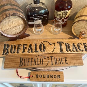 Puede incluir: Se muestran letreros de madera y una etiqueta con el texto "Buffalo Trace" y "Bourbon". Tambi&eacute;n se ven dos botellas de bourbon y dos vasos, junto con dos peque&ntilde;os barriles de madera.