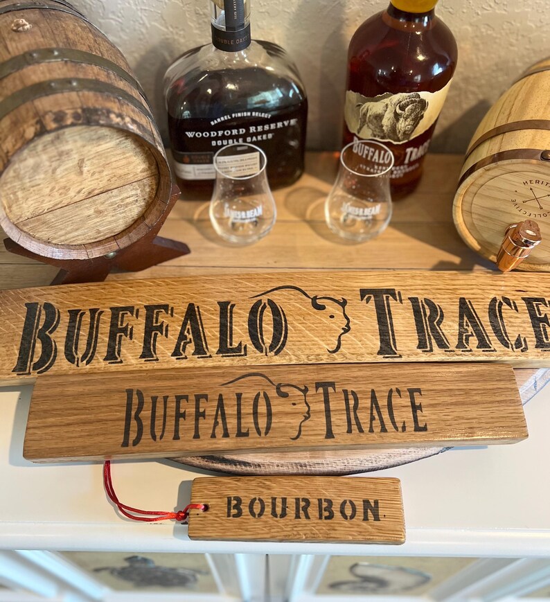 Puede incluir: Se muestran letreros de madera y una etiqueta con el texto "Buffalo Trace" y "Bourbon". Tambi&eacute;n se ven dos botellas de bourbon y dos vasos, junto con dos peque&ntilde;os barriles de madera.
