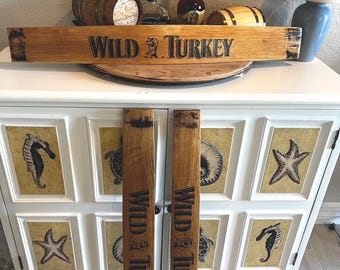 May include: Wooden Wild Turkey sign and barrel stave pieces. The sign is rectangular with the words "WILD TURKEY" in black lettering. Two barrel staves are also visible, each with the same text. The items are displayed on a white cabinet.