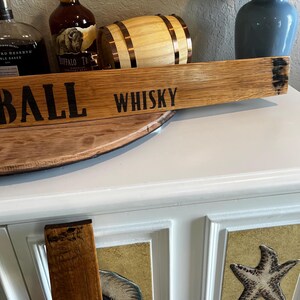 Puede incluir: Un letrero de madera con la palabra "WHISKY" en letras negras. El letrero est&aacute; hecho de un trozo de barril y tiene un aspecto r&uacute;stico. Otros elementos en la imagen incluyen un peque&ntilde;o barril de madera y botellas de whisky.